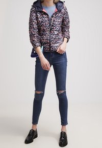 Giacca imbottita a fantasia floreale in navy con chiusura lampo, texture ondulata e cappuccio. Abbinata a jeans skinny blu e scarpe nere.
