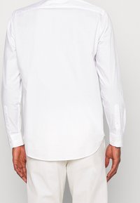 Man in een witte overhemd met lange mouwen en witte broek, van achteren gezien tegen een effen achtergrond.