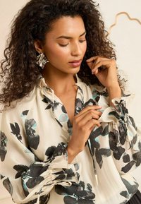 Floral blouse in ivory with black and turquoise patterns, featuring elasticized cuffs and ties at the neckline, made of lightweight fabric.