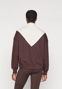 Brun genser med en hvit skråpanel. Har strikket tekstur og ribbestrikkede mansjetter. Båret over matchende brune leggings.