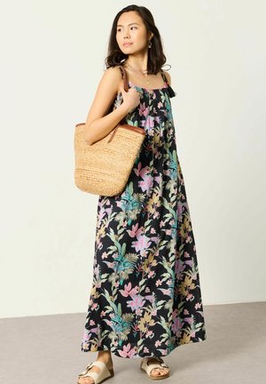 Frau in einem ärmellosen schwarzen Maxikleid mit Blumenmuster, die eine geflochtene Strohtasche hält und beige Sandalen trägt, schaut zur Seite.