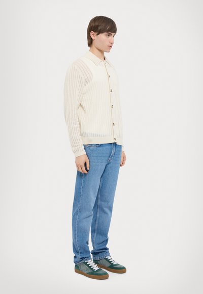 Light cream ribbed cardigan with buttons, paired with blue jeans and green-accented sneakers. Simple, textured design, fitted silhouette.