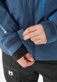 Person adjusting blue Gore-Tex jacket cuff with Velcro strap, wearing black Millet pants with logo on front pocket.