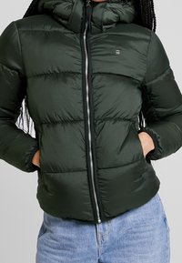 Svart-grön vadderad pufferjacka med framficka, huva och sidofickor, bärs tillsammans med ljusblå denimjeans.