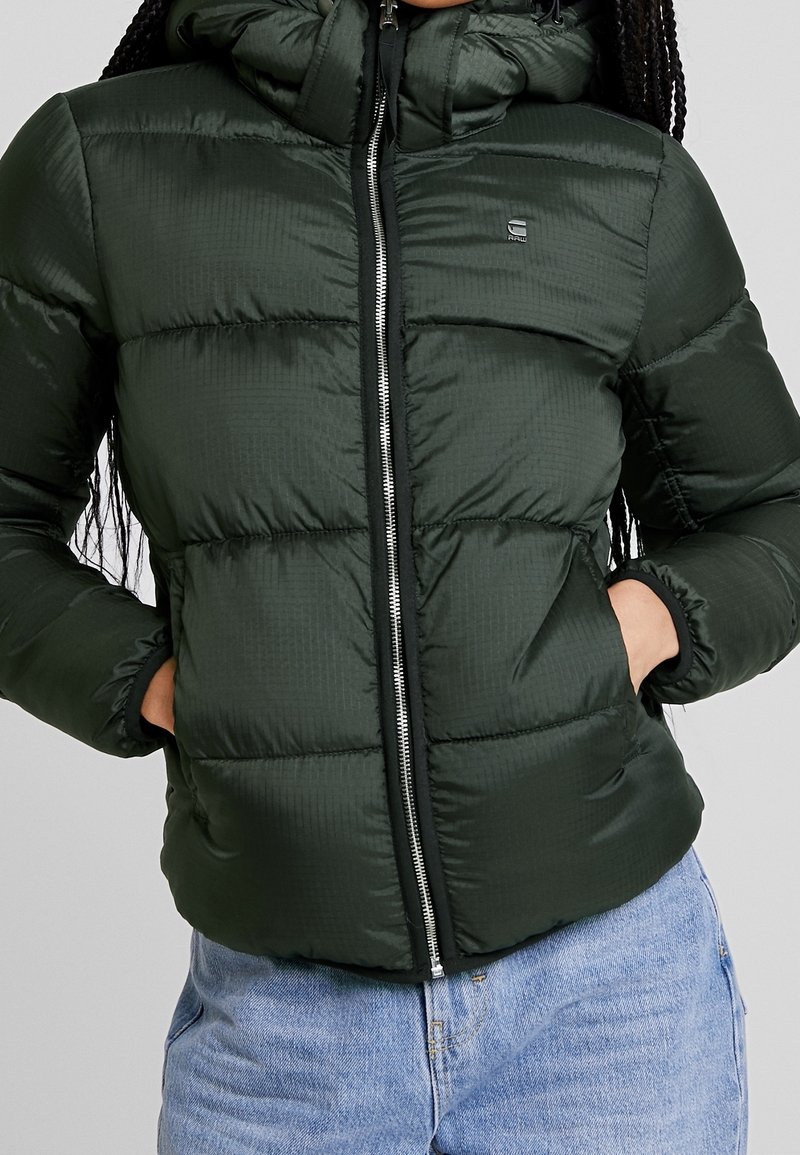 Svart-grön vadderad pufferjacka med framficka, huva och sidofickor, bärs tillsammans med ljusblå denimjeans.