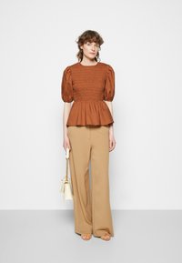 Top peplum castanho com detalhes franzidos e mangas bufantes, combinado com calças largas bege, segurando uma mala creme. Textura de tecido suave.