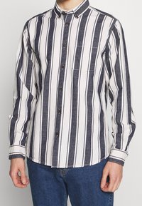 Camicia button-up con righe verticali blu navy e bianche, una tasca sul petto e bottoni marroni a contrasto. Realizzata in tessuto intrecciato con finitura texture.