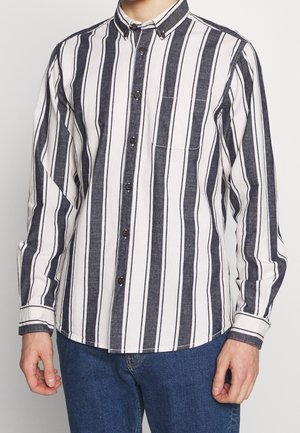 Skjorta med knappar framtill som har vertikala marinblå och vita ränder, en bröstficka och kontrasterande bruna knappar. Tillverkad av vävd tyg med en texturerad finish.