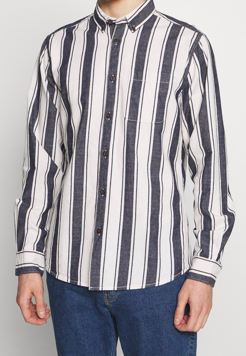 Camicia button-up con righe verticali blu navy e bianche, una tasca sul petto e bottoni marroni a contrasto. Realizzata in tessuto intrecciato con finitura texture.