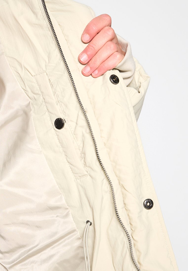 Helle Jacke aus strukturiertem Stoff, mit Reißverschluss, Druckknöpfen und Kordelzugdetails. Zeigt Innenfutter und Tasche.