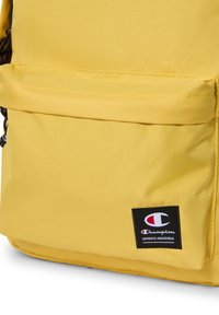 Żółty plecak z tkaniny o teksturowanej powierzchni, z przednią kieszenią i czarną naszywką z logo Champion. Prosty, funkcjonalny design.