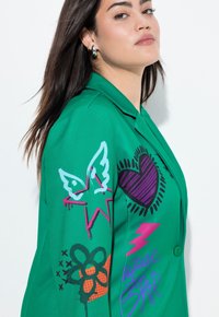 Groene blazer met een oversized pasvorm, met kleurrijke graphics waaronder harten, vlinders en bliksemschichten. Soepele stof met knoopsluiting.