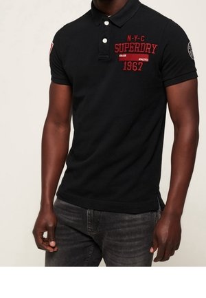 Mand iført en sort Superdry polo med rød tekst "N-Y-C Superdry 1962" og mørkegrå jeans foran en ensartet lys baggrund.