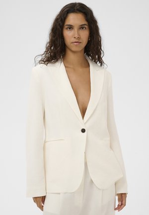 Femeie cu păr creț și închis la culoare, purtând un blazer crem cu un singur nasture și decolteu deschis, pe un fundal simplu, deschis.