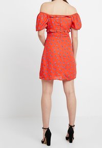 Červené šaty bez ramínek s nabíranými rukávy, květinovým vzorem, smockovaným pasem a rozšířenou sukní. Doplněné černými sandály na vysokém podpatku.