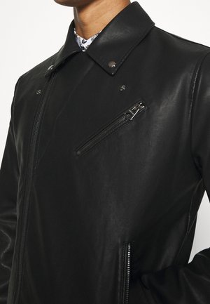 Man draagt een zwarte leren jas met ritsen op de borst- en zijzakken, en een kraag met zilveren drukknopen over een geblokt overhemd.