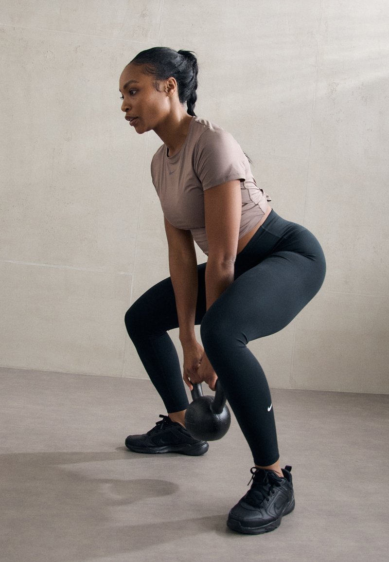 Kettlebell nera tenuta da una persona in una maglietta marrone chiaro e leggings neri, su uno sfondo di parete e pavimento dai toni neutri.