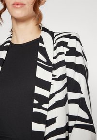 Sako s černobílým pruhovaným vzorem zebra, hladkou texturou a otevřeným designem vpředu, doplněné úzkým černým topem s ribbovým vzorem.