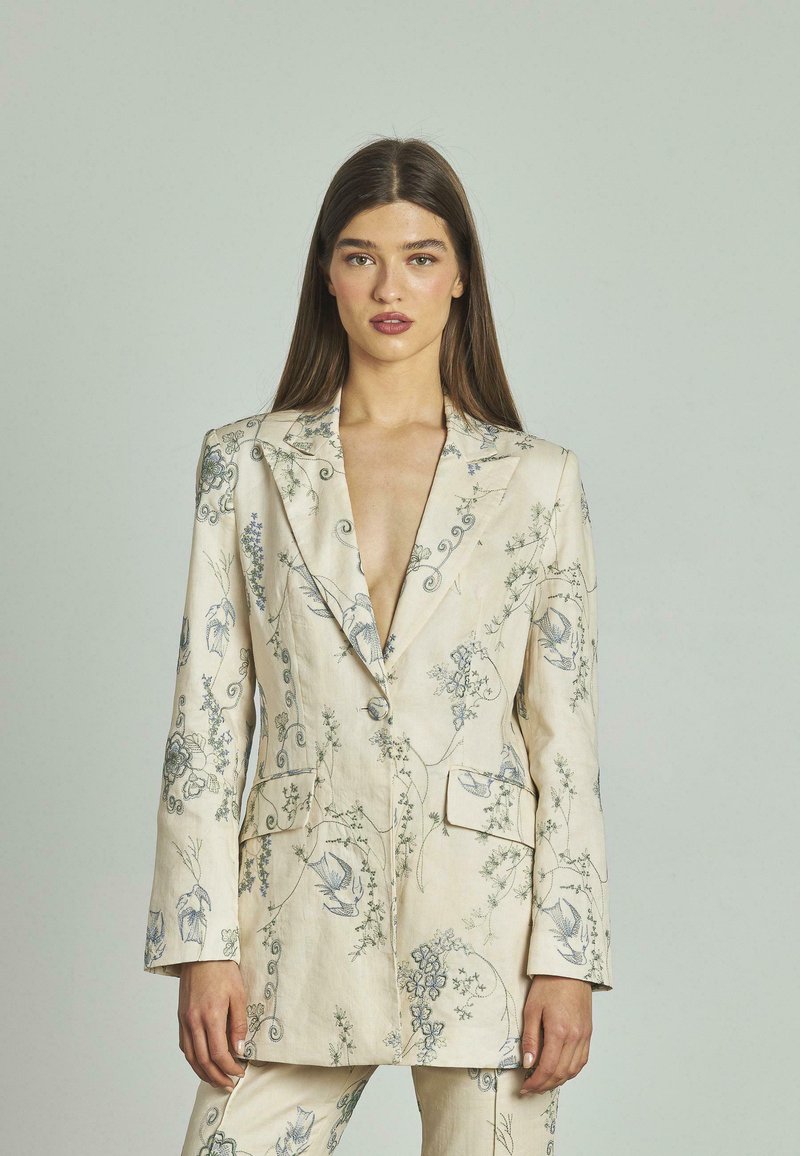 Blazer in lino beige con dettagli di ricamo floreale blu, scollatura a V profonda, rever, e due tasche frontali.