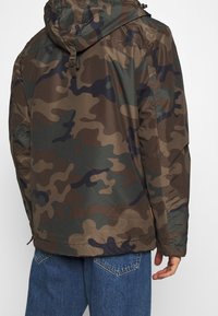 Maastokuvioinen takki ruskean, vihreän ja tummansinisen sävyissä; mukana hupullinen, teksturoitu kangas ja säädettävä helma. Takakatselu esitetty.