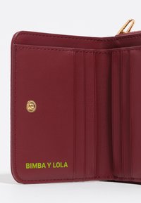 Cartera Bimba y Lola en cuero burdeos, con compartimentos para tarjetas, herrajes dorados y un logo verde en la esquina inferior derecha.