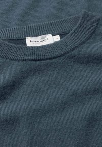 Maglione lavorato a maglia di un verde scuro con scollo tondo a costine. La trama del tessuto è morbida e liscia. L'etichetta "hessnatur" è visibile vicino al colletto.