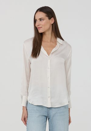 Camicia a maniche lunghe color beige chiaro realizzata in tessuto morbido, con un'apertura frontale con bottoni, colletto classico e orlo leggermente curvo.