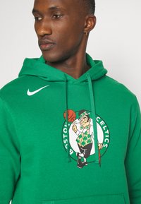 Zelená mikina vyrobená z bavlneného zmesi, s centrálnym logom Boston Celtics, na ktorom je zobrazený leprikón a basketbal. Obsahuje šnúrku na stiahnutie a prednú kapsu.
