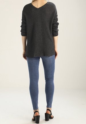 Maglione grigio a maglia con scollo a V, maniche arrotolate e un retro leggermente più lungo. Abbinato a jeans skinny blu e sandali neri con tacco.