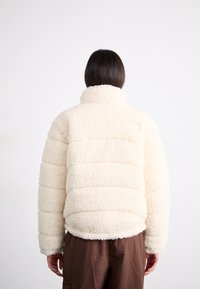 Jasno kremowa polarowa kurtka o puchowym, pikowanym wzorze, z wysokim kołnierzem i elastycznym dołem. Tył kurtki zdobi małe logo. Teksturowana, miękka powierzchnia.