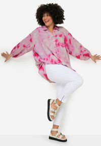 Blusa leggera in tie-dye rosa e crema, con bottone frontale e maniche a sbuffo. Abbinata a pantaloni bianchi e sandali con laccetti.
