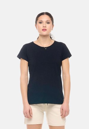 Jeune femme aux cheveux foncés attachés en arrière, portant un t-shirt noir à manches courtes et un short beige, debout devant un fond clair uni.