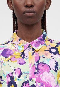 Close-up van een persoon met gevlochten haar die een gebuttond gebloemd shirt draagt met paarse, gele en pastelkleurige tinten op een lichte achtergrond.