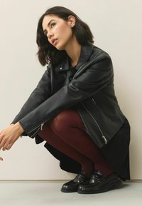 Schwarze Leder-Motorradjacke mit Reißverschlüssen. Das Modell trägt kastanienbraune Leggings und schwarze Slipper. Glatte Textur, taillierter Schnitt, minimalistische Details.