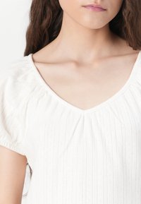 Bílá blůzka s výstřihem do V, krátkými nabíranými rukávy a vertikálním žebrovaným vzorem se spoustou malých diamantových motivů.