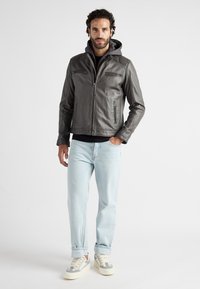 Grijze leren herenjack met een capuchon, voorzien van ritszakken aan de voorkant, in een rechte pasvorm, gedragen met lichtblauwe jeans en sneakers.