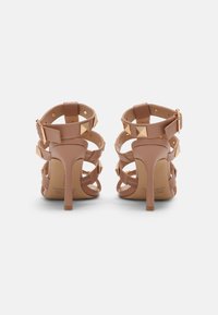 Paire de sandales à talons hauts nude avec plusieurs brides et clous pyramidaux dorés, vues de dos sur fond blanc.