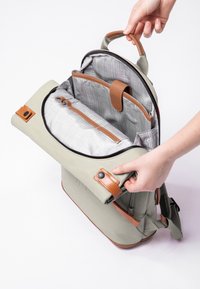 Zaino grigio chiaro con dettagli in pelle marrone, una maniglia superiore e un ampio interno caratterizzato da un motivo a quadretti e diversi scomparti.