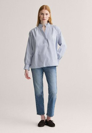 Kvinde iført en blå- og hvidstribet bluse med flæsekant ved krave og manchetter, blå jeans og mørkebrune mokkassinsko, stående.