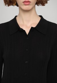 Gros plan d'une femme portant une chemise noire côtelée à boutons avec un large col et des cheveux bouclés rouges.