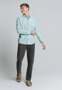 Lichtblauw overhemd met knopen en donkergrijze broek, aangevuld met bruine sandalen. Het overhemd heeft een klassieke kraag en lange mouwen.