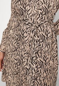 Beige klänning med svart abstrakt tigerstrimmönster, med knytmidja, knapprad och långa ärmar. Tillverkad av mjukt tyg.