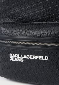 Mochila preta com padrão KL em relevo, fecho metálico e texto logo em branco a dizer "KARL LAGERFELD JEANS." Material texturizado suave.