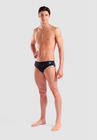 Svarta badbyxor med blå logga, åtsittande design, slät textur och låg midja, visade av en manlig modell mot en neutral bakgrund.