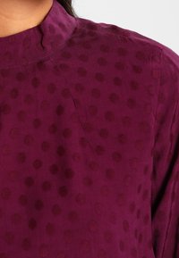 Blouse violette texturée avec un col montant, présentant un motif de points en relief d'une teinte plus foncée, mettant en valeur un tissu doux.