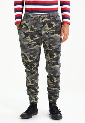 Man draagt groene en zwarte camouflage joggingbroek met zwarte sneakers en een rood-wit-blauw gestreept shirt met lange mouwen.