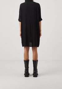 Abito camicia nero oversize con maniche corte arrotolate, tessuto liscio e orlo leggermente curvato, abbinato a stivali neri arrivanti fino al ginocchio.