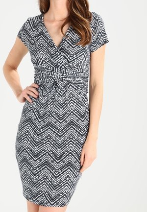 Kobieta w czarno-białej, dopasowanej sukience z krótkim rękawem i wzorem w kropeczki w kształcie chevron, pozująca z jedną ręką na biodrze, a drugą swobodnie opuszczoną.