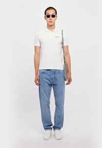 Hvit polo skjorte med logo, lyseblå jeans, hvite joggesko, grå skulderveske og svarte solbriller. Modell som står mot en ensfarget bakgrunn.