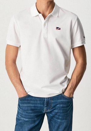 Hombre vistiendo una camisa polo blanca de manga corta con un pequeño logo de bandera en el pecho y jeans azules, con las manos en los bolsillos, mirando de frente.